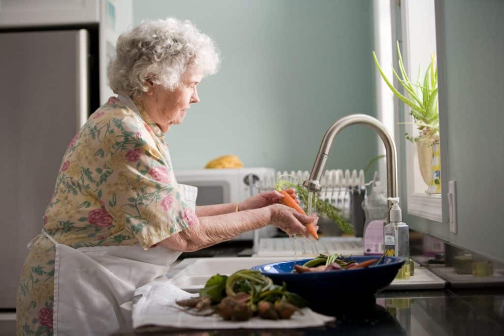 Femme âgée avec un tablier en train de laver ses légumes dans l'évier de sa maison.